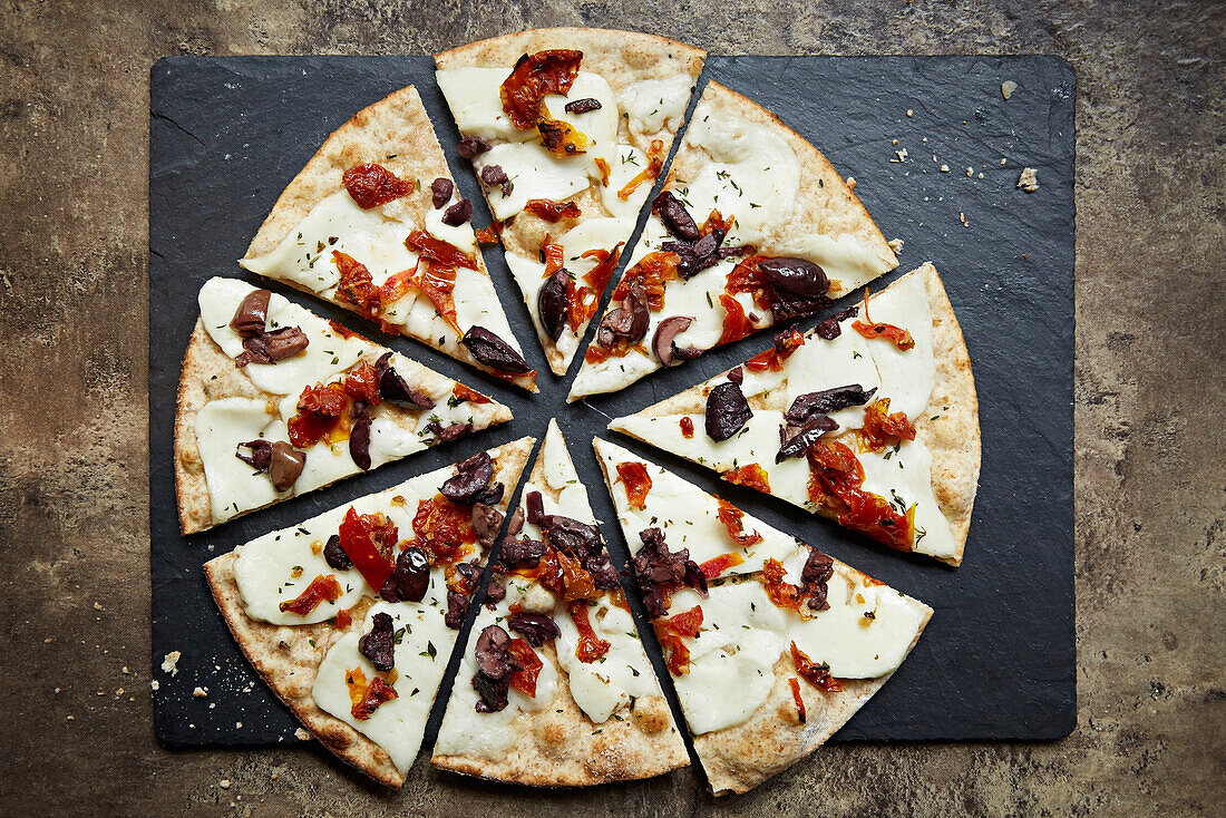 A pizza topped with halloumi, black olives and peppers, sliced on a slate platter