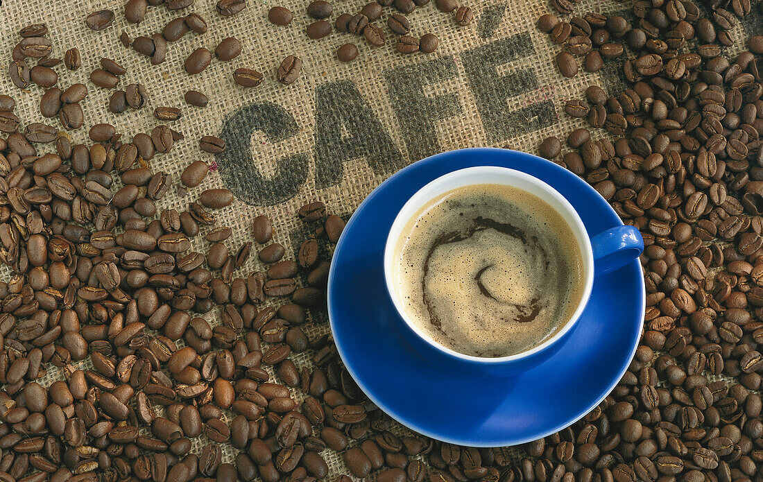 A blue cup of coffee surrounded by coffee beans on a coffee bag labelled 'Cafe'.