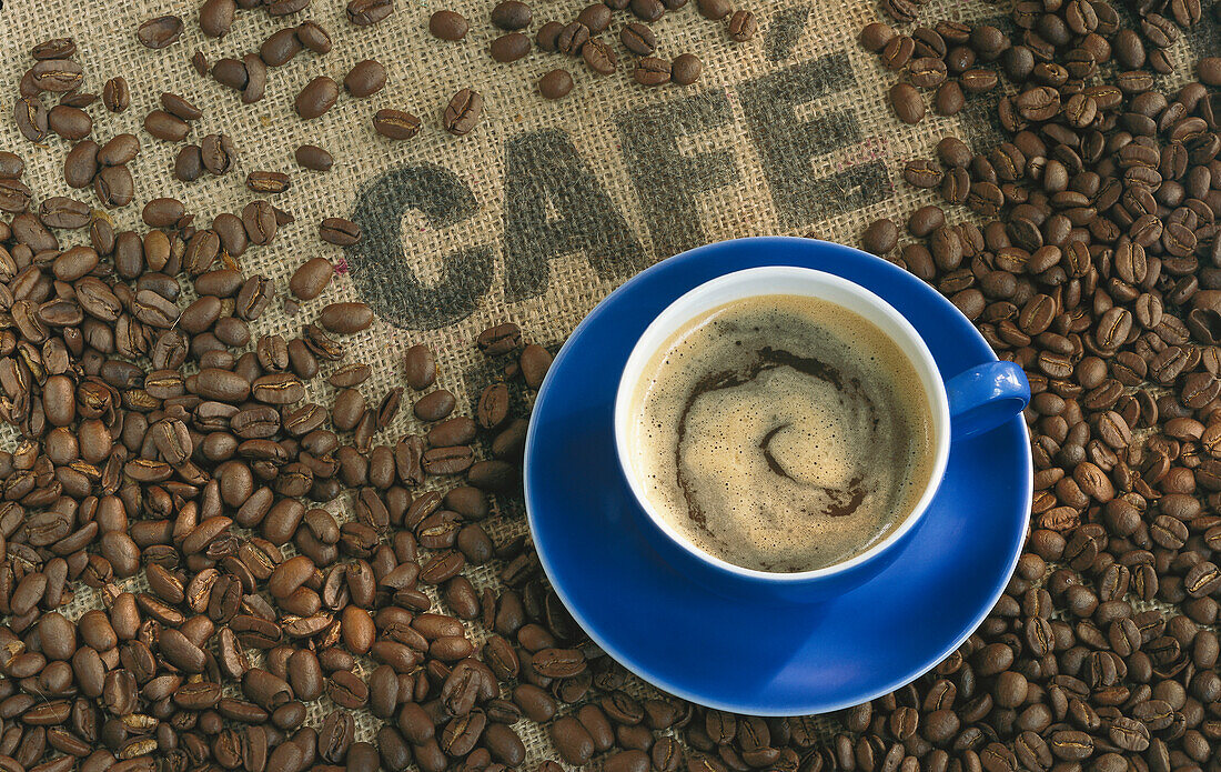 Eine blaue Tasse mit Kaffee, umgeben von Kaffeebohnen auf einem Kaffeesack mit Aufschrift 'Cafe'