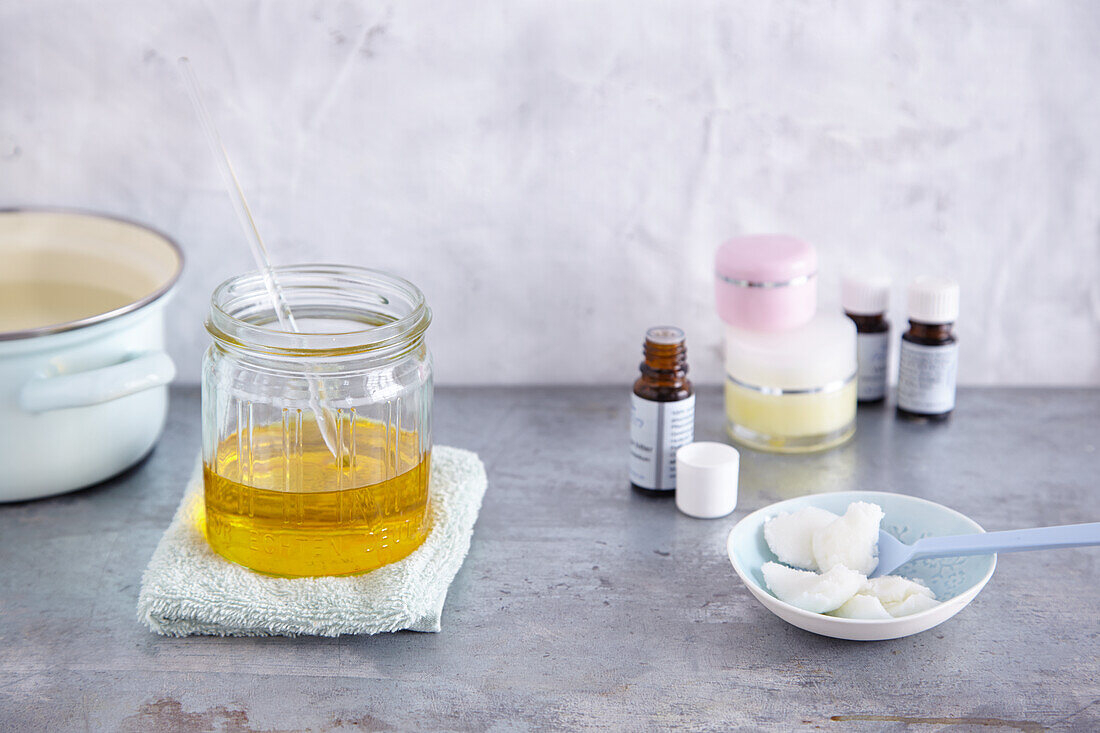 Glass jar with base oil, coconut oil in a small bowl (coconut oil should be melted in the oil), jar and essential oil bottles in the background