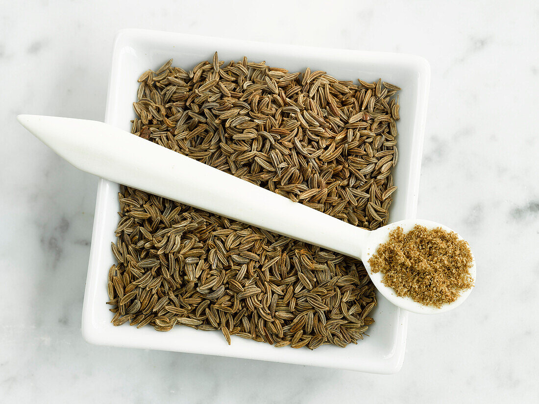 A bowl with cumin and a spoon with cumin powder on a light background