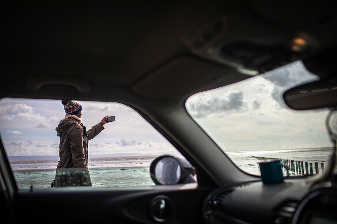 Man with camera phone on winter beach outside car