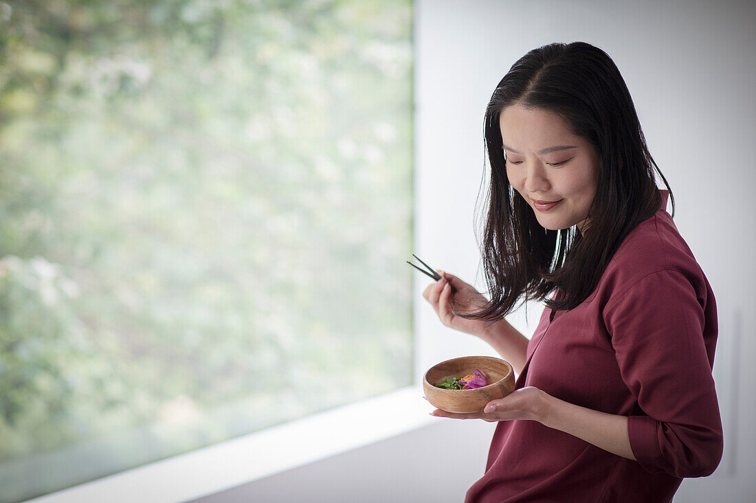 Young woman eating from bowl with chopsticks at window