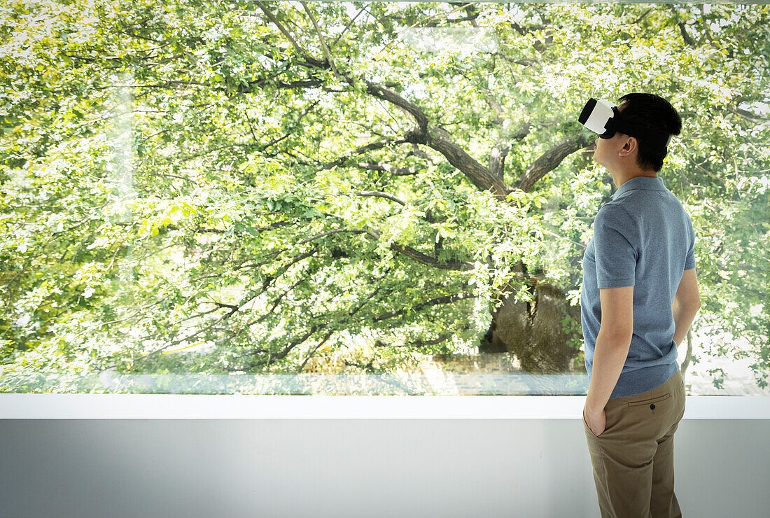Man using VR headset at window with view of green tree