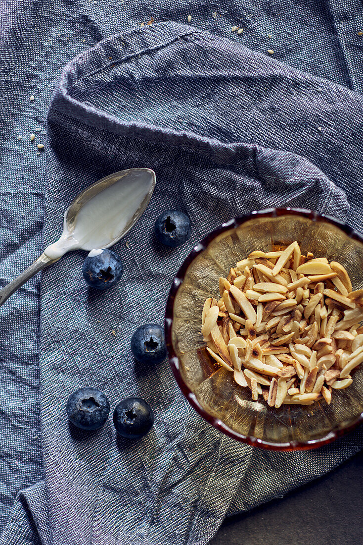 Roasted almonds in a small bowl next to blueberries and a spoon covered in yoghurt