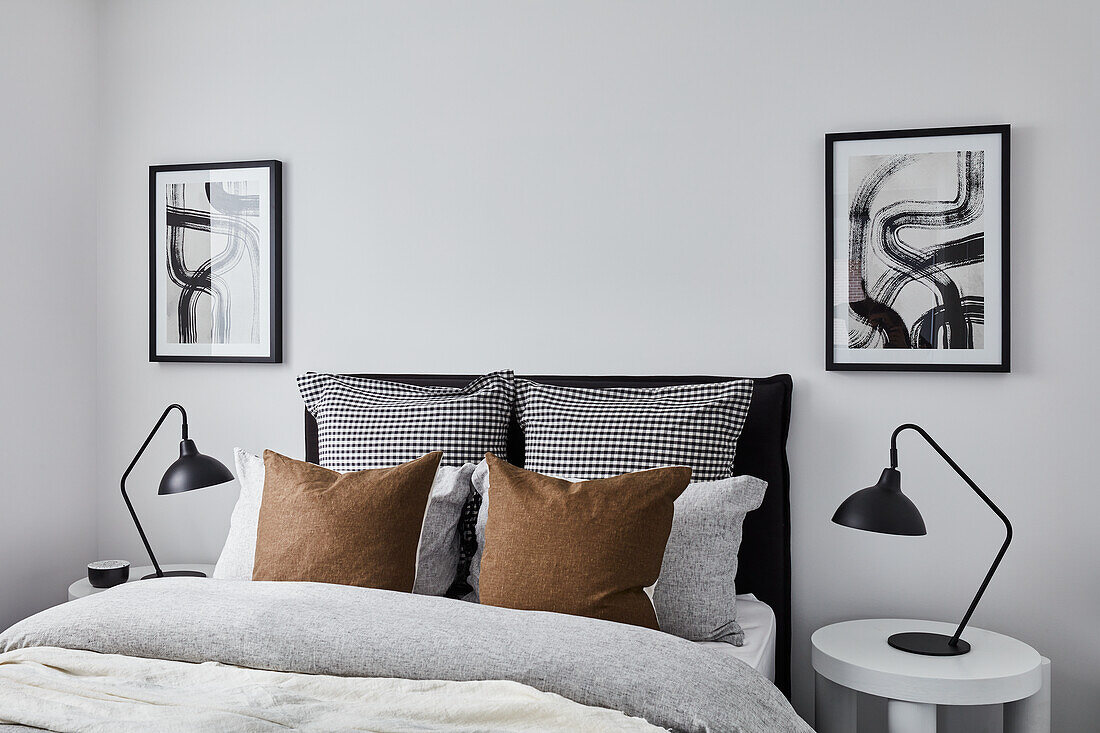 A modern monochromatic bedroom with grey and white linen bedding and light-brown cushions, abstract art on the wall, white side tables and black lamps