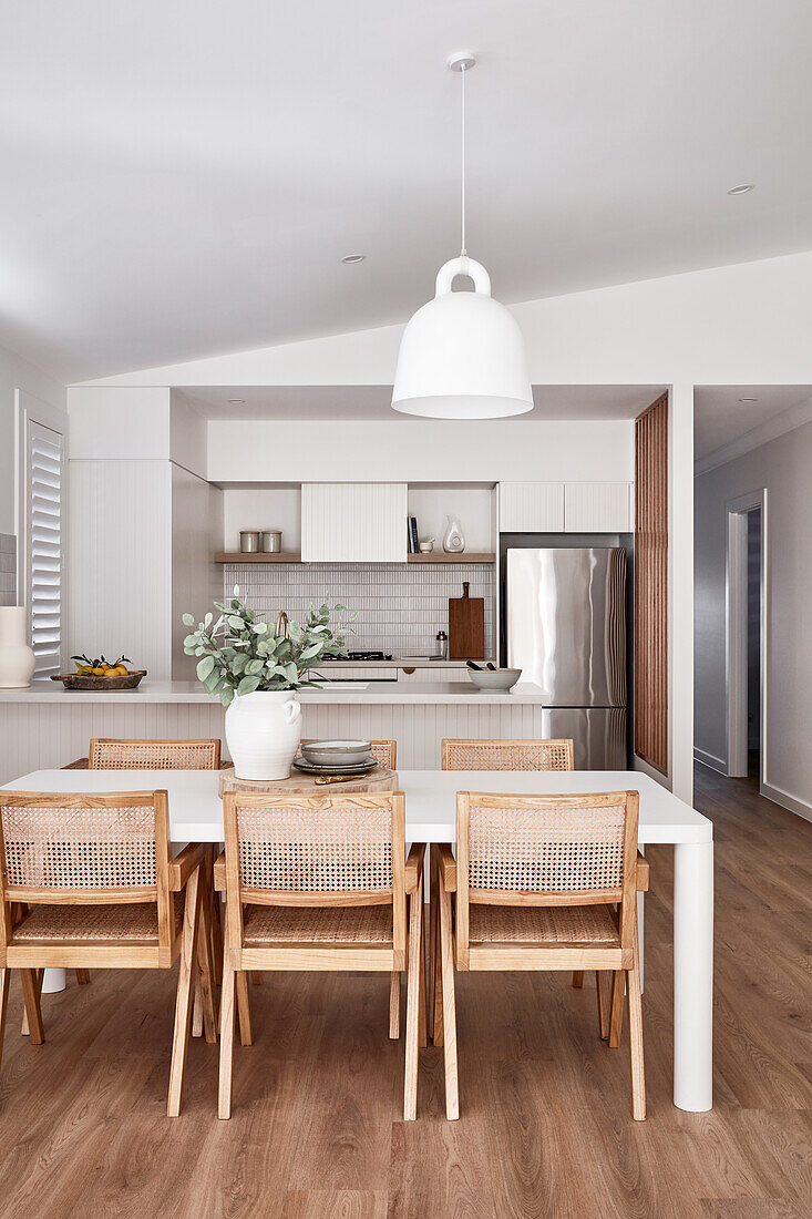 An open-plan kitchen and a dining room in a modern Scandi-style with white furniture and woven chairs and an oak wood floor