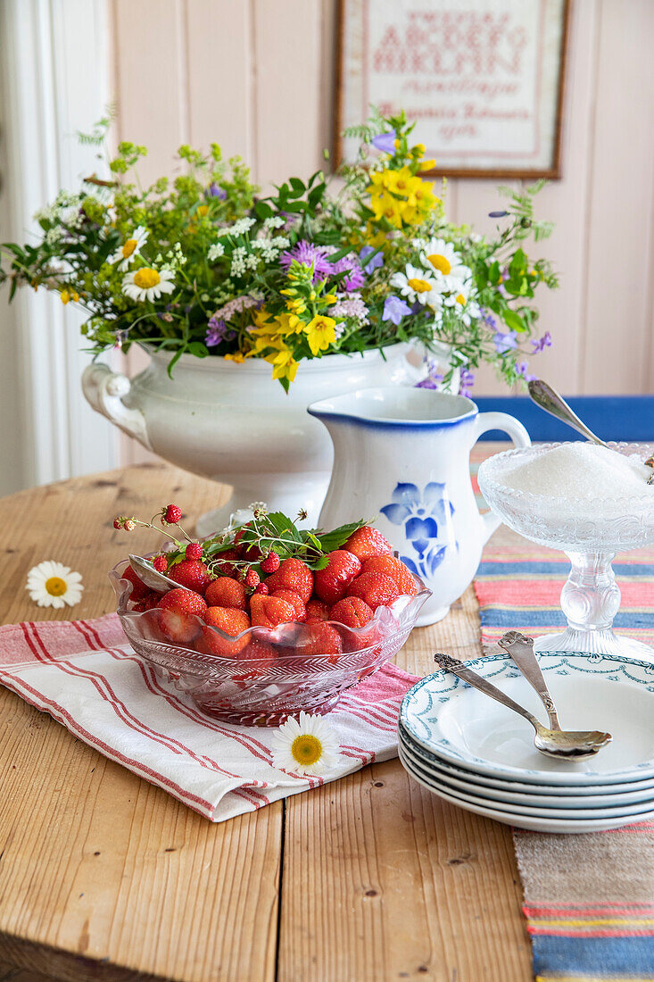 Glass bowl with strawberries, plate, pitcher, and bouquet of flowers on a kitchen table