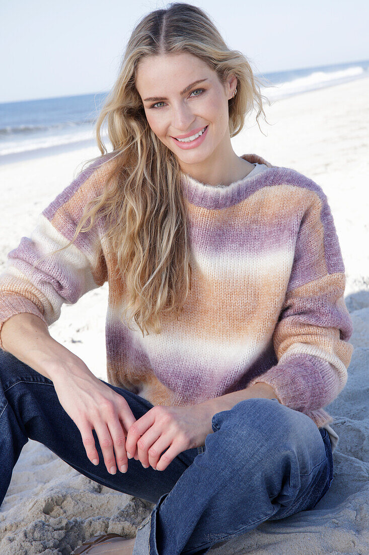 Junge blonde Frau im Strickpullover mit Farbverlauf und Jeans am Meer