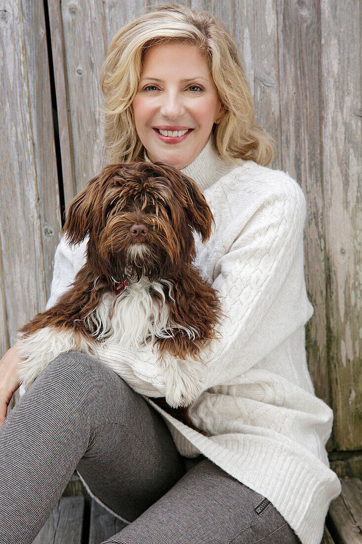 A mature blonde woman holding a dog wearing a white knitted jumper and grey leggings