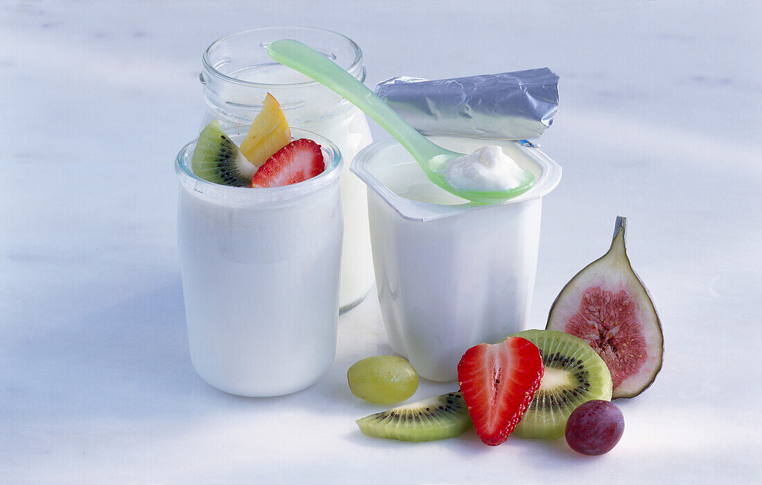 Ein Becher Joghurt mit Löffel und zwei Gläser Joghurt davor Obst
