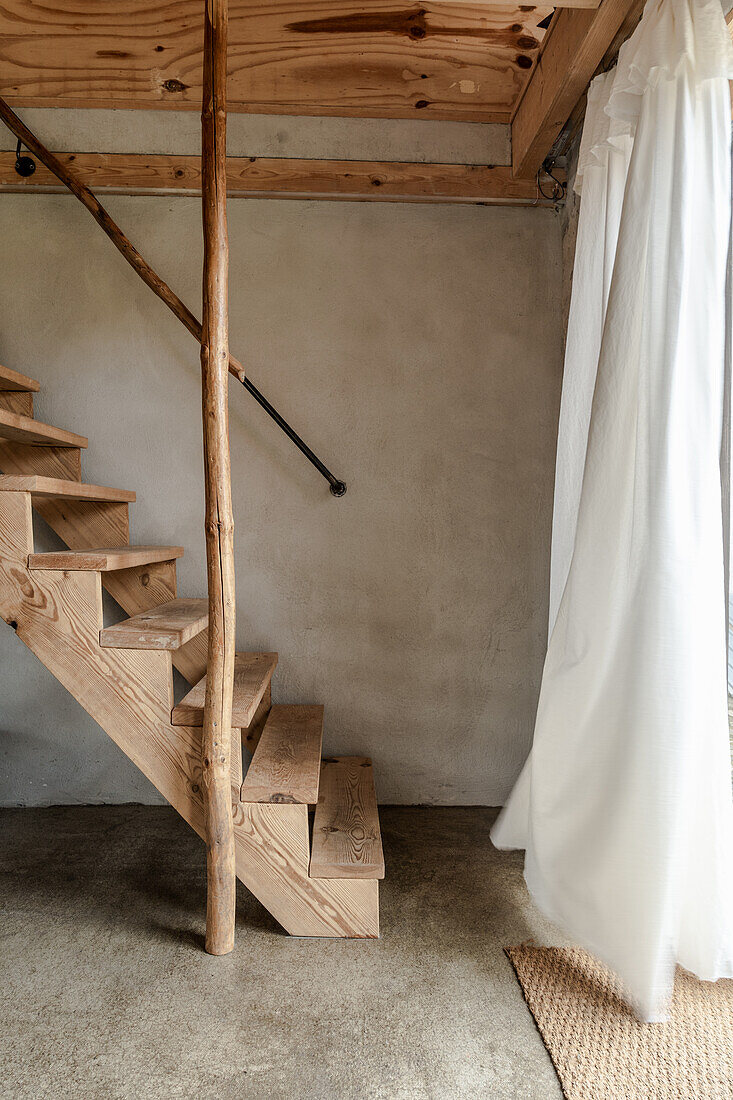 Holztreppe im Raum mit Estrichboden, weißer wehender Vorhang vor Fenster