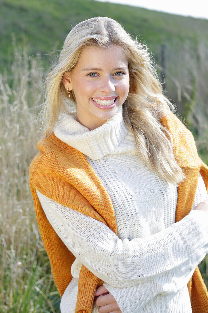 Junge blonde Frau in weißem Rollkragenpullover, mit gelbem Strickpulli über den Schultern in der Natur