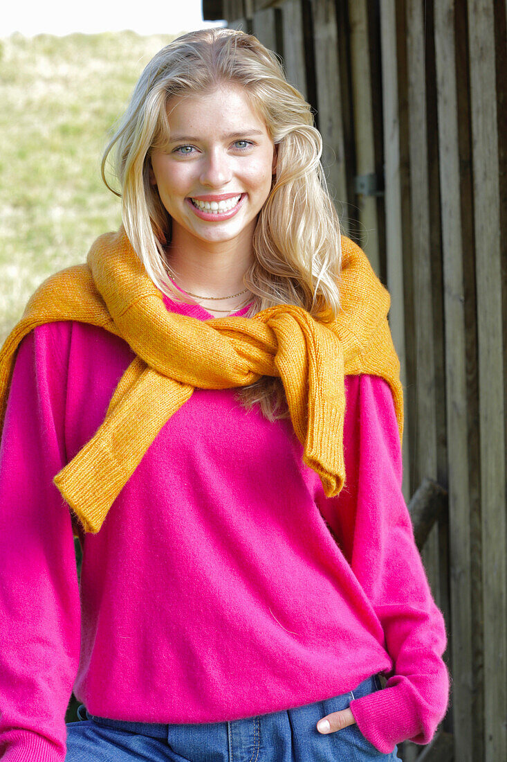Junge blonde Frau in pinkfarbenem Pullover, mit gelbem Strickpulli über den Schultern im Freien