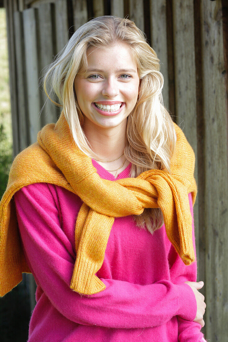 Young blonde woman in a pink sweater with a yellow knit sweater over her shoulders in front of a wooden wall
