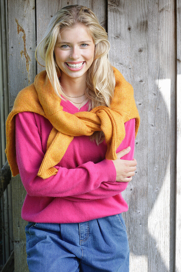 Junge blonde Frau in pinkfarbenem Pullover, mit gelbem Strickpulli über den Schultern vor Bretterwand