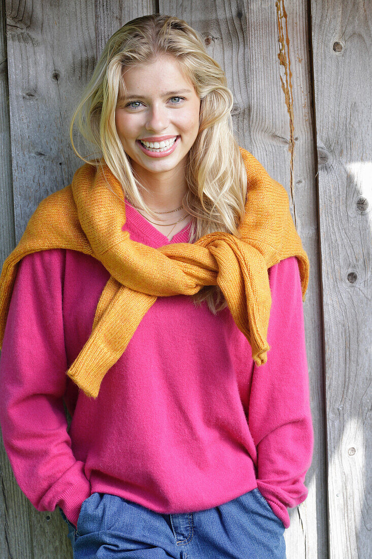 Junge blonde Frau in pinkfarbenem Pullover, mit gelbem Strickpulli über den Schultern vor Bretterwand