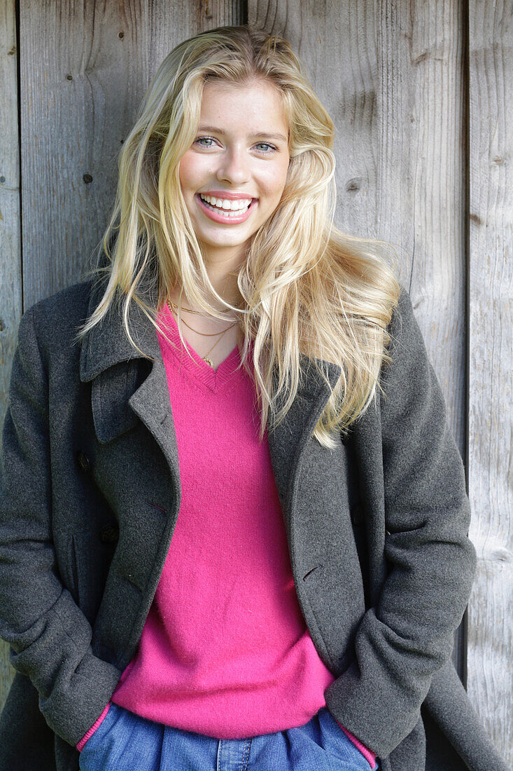 Young blonde woman in a pink sweater and gray wool coat in front of a wooden wall