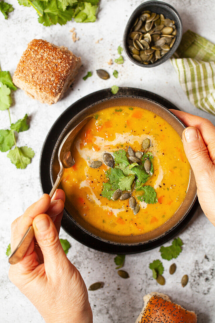 A person eating a bowl of carrot soup that has been garnished with fresh coriander and pumpkin seeds