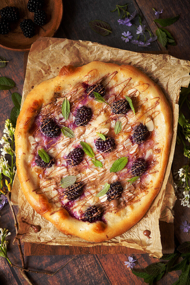 A sweet dessert pizza topped with blackberries, chocolate drizzle, almonds and mint
