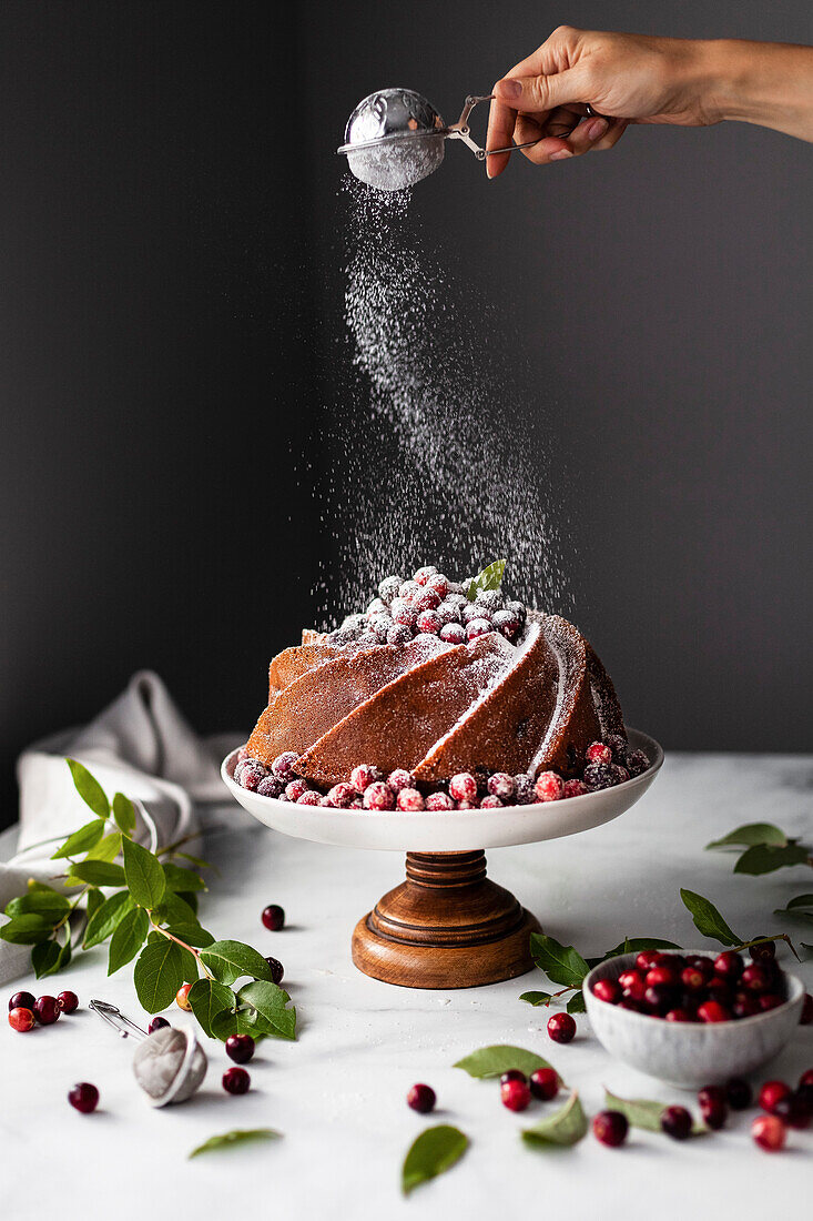 A baker dusting a cranberry orange bundt cake with powdered sugar