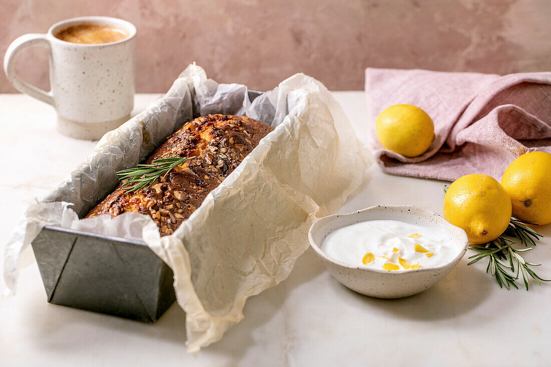 Homemade lemon cake with nuts and rosemary in baking dish, serving with fresh lemon, white sauce and coffee mug on white marble table