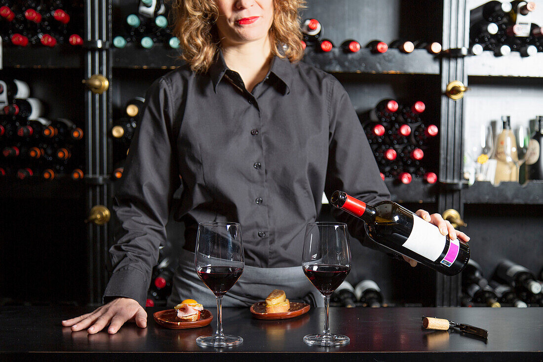Crop anonymous female barkeeper pouring red wine into wineglass while standing at counter with served foie gras during work in restaurant