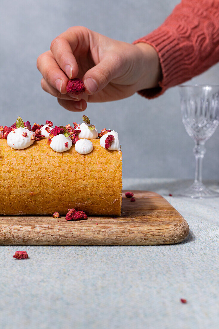 Crop anonymous female topping sweet roll cake with dried raspberries on wooden board near glass goblet