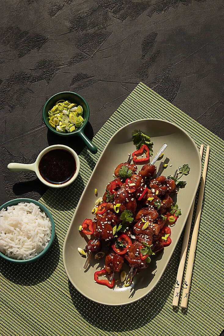 Top view of delicious marinated chicken on skewers topped with greens served on table with bowls of rice, soy sauce and leek in restaurant