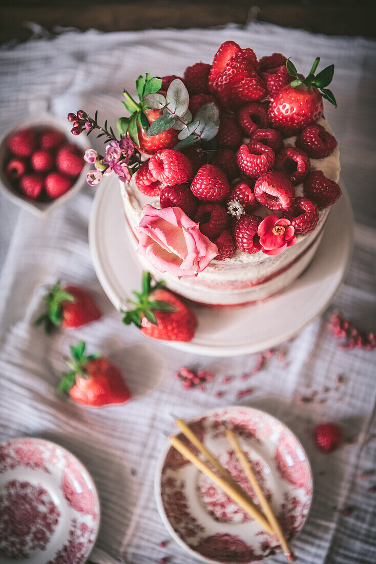 From above delicious red velvet cake topped with fresh strawberries and decorated with flowers served on table with white tablecloth in kitchen