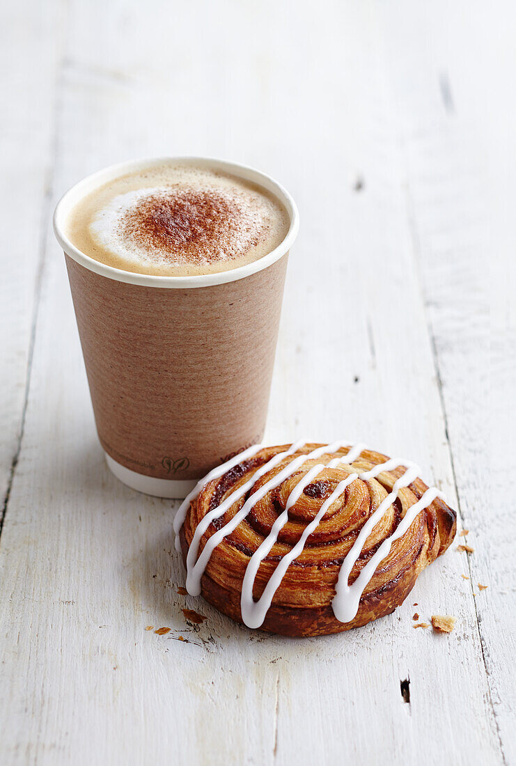Frothy takeaway cappuccino next to a cinnamon swirl pastry drizzled with icing
