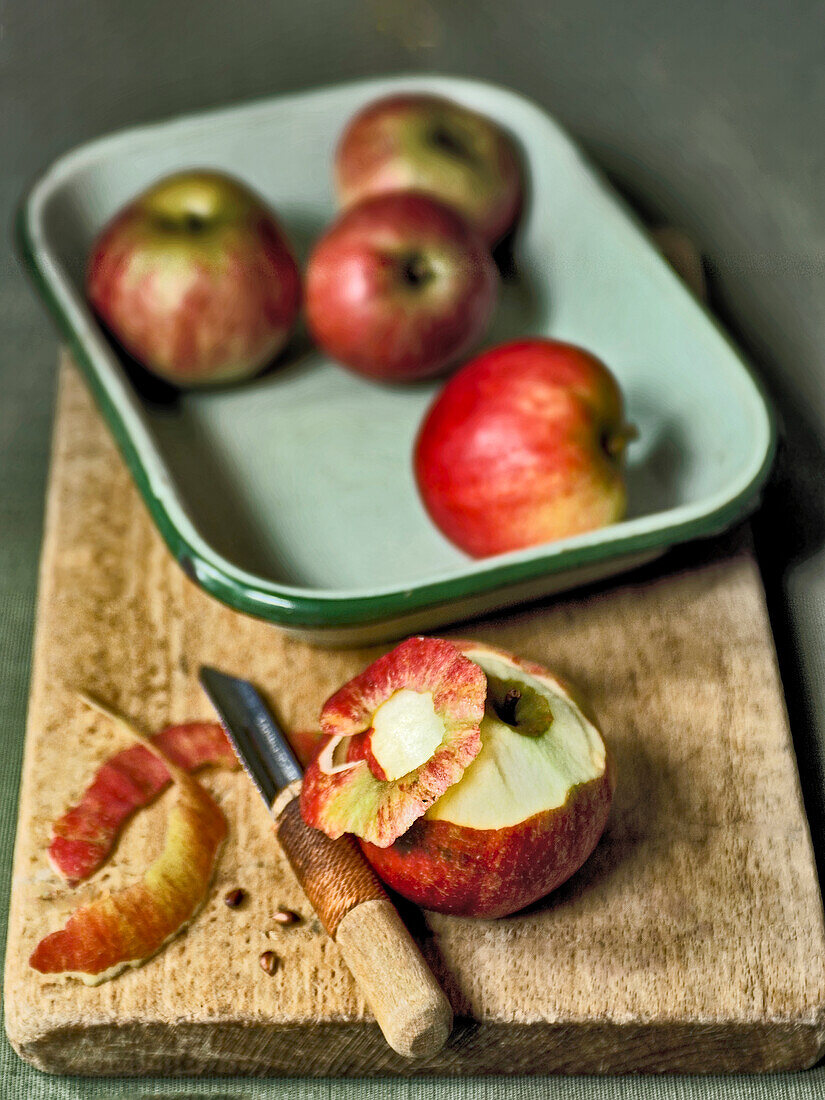 Unpeeled apples in an enamel dish and a partially peeled apple with a peeler on a wooden cutting board