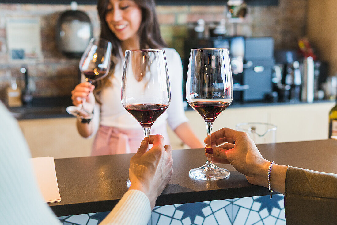 Female bartender in smart casual clothes enjoying aroma of red wine while sitting at counter during degustation session in bar with cropped unrecognizable female sommeliers
