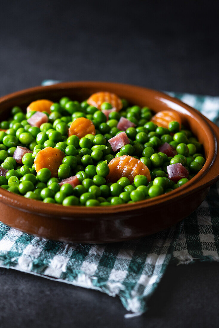 Bowl of delicious green peas with ham and carrot placed on napkin on dark background