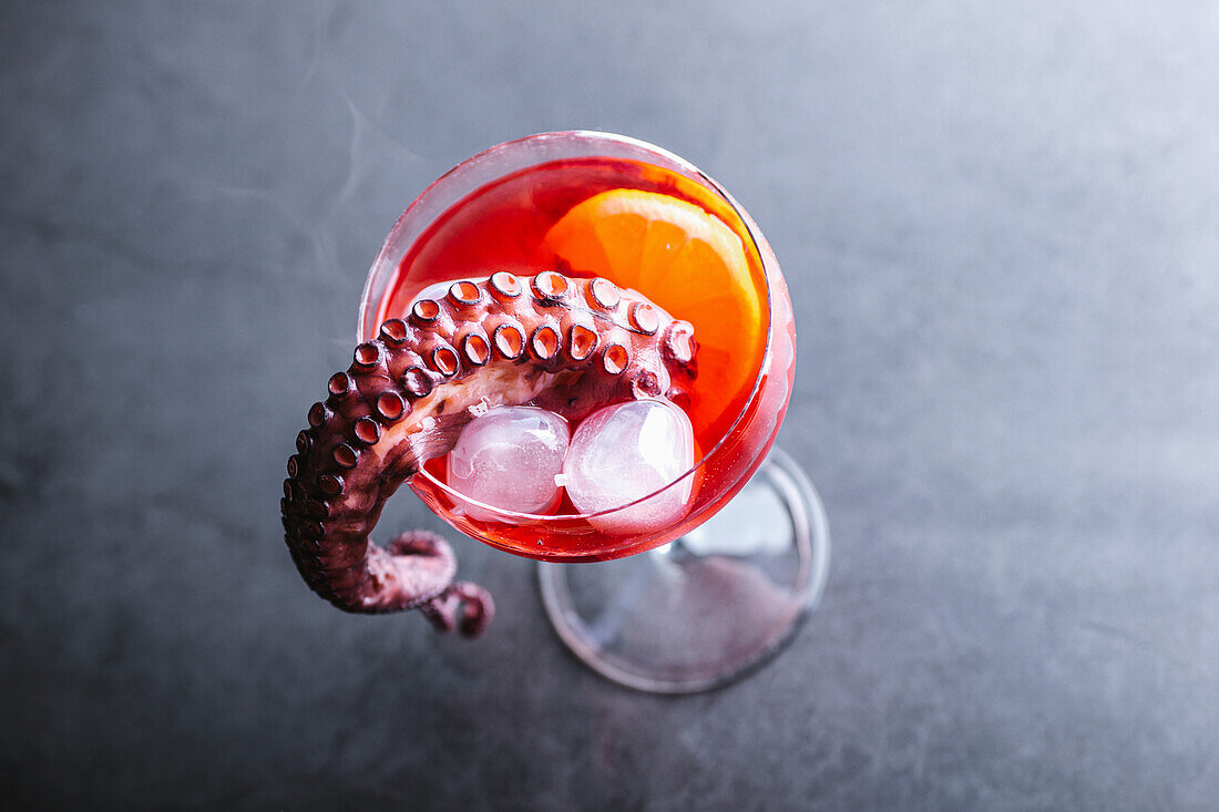 From above alcoholic cocktail with ice cubes served with octopus tentacle and lemon slice placed on table against gray background in studio