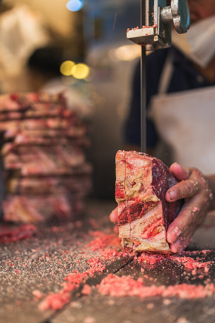 Anonymous mature man in apron and face mask using electric tabletop saw to slice piece of frozen meat during work in butchery