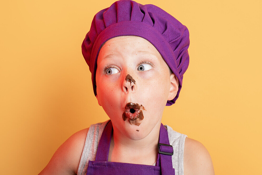 Astonished messy boy in purple chef uniform with sweet chocolate on mouth looking away while standing on yellow background in studio