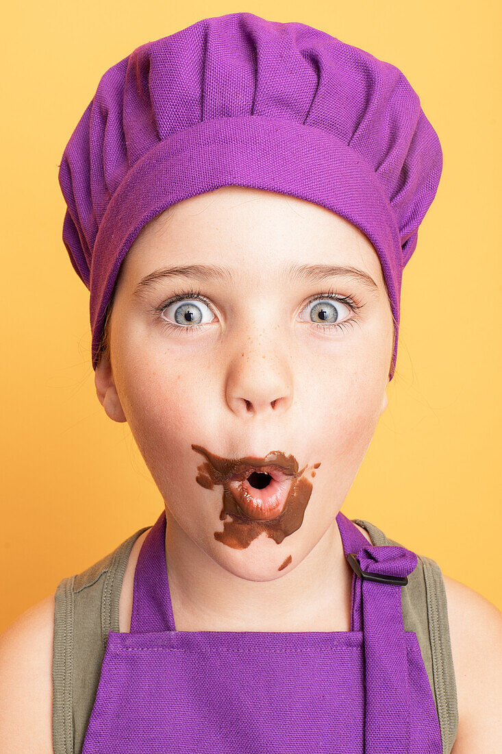 Astonished messy girl in purple chef uniform with sweet chocolate on mouth looking at camera while standing on yellow background in studio