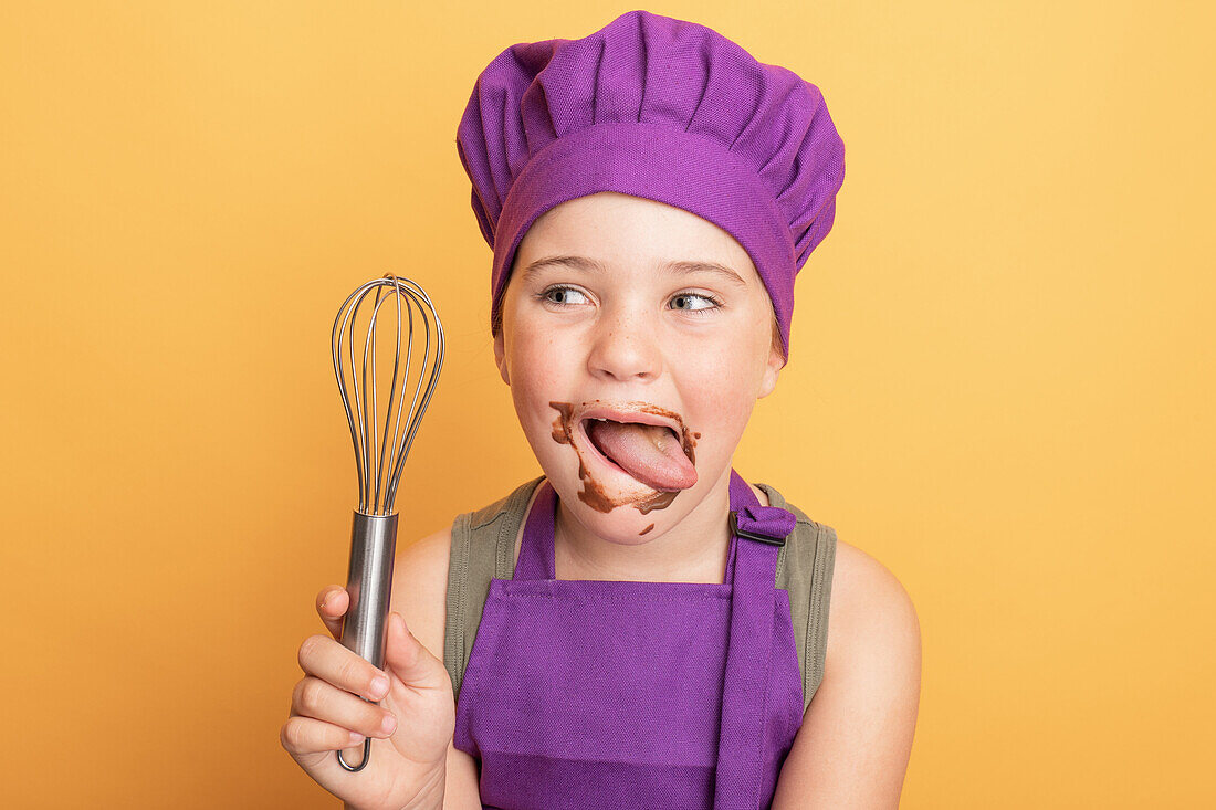 Cute smiling dirty boy in chef uniform looking at camera holding whisk while licking sweet chocolate off face against yellow background in studio