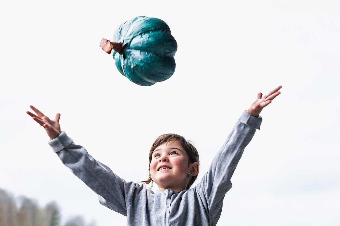 Positive girl in casual wear throwing teal pumpkin in air with raised arms while standing in countryside against cloudless sky