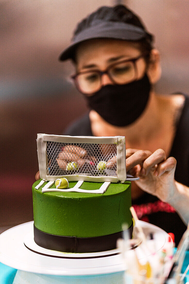 SWoman in face mask arranging sweet tennis balls near net on fondant cake during work in bakery