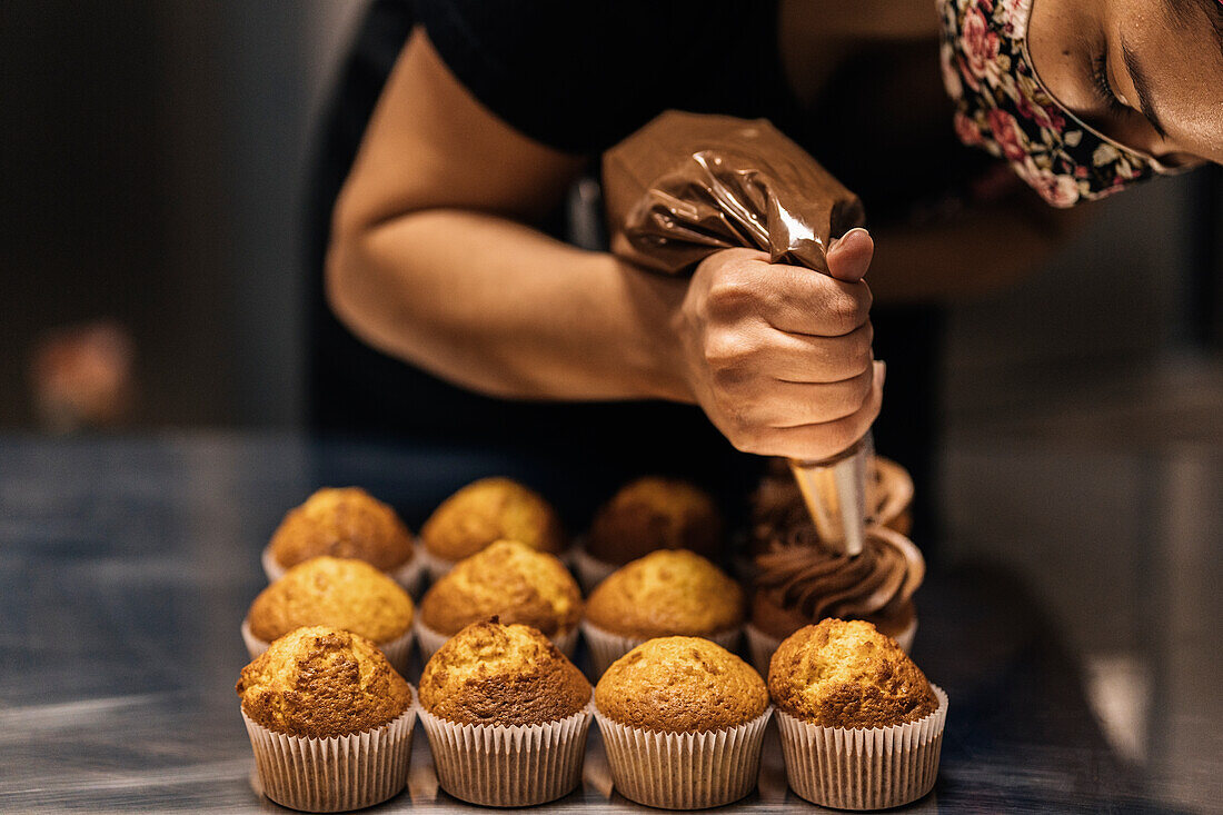 Frau mit Gesichtsmaske verziert Cupcakes mit Schokoladencreme in einer Konditorei