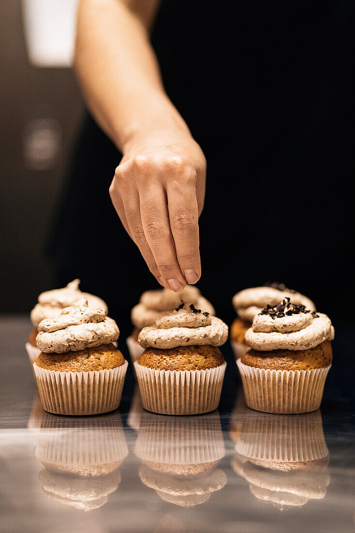 Anonymous female baker scattering chocolate sprinkles on top of delicious muffins during work in bakehouse