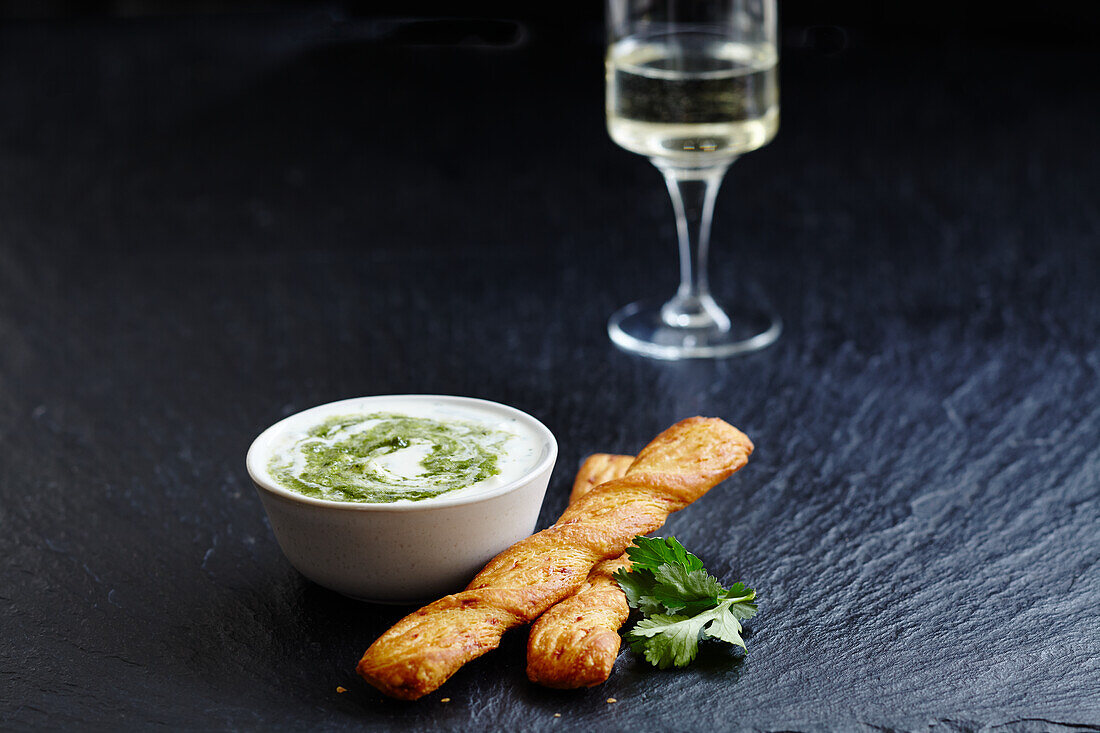 Käsestangen mit Pesto-Dip dazu ein Glas Weißwein auf schwarzem Schieferuntergrund