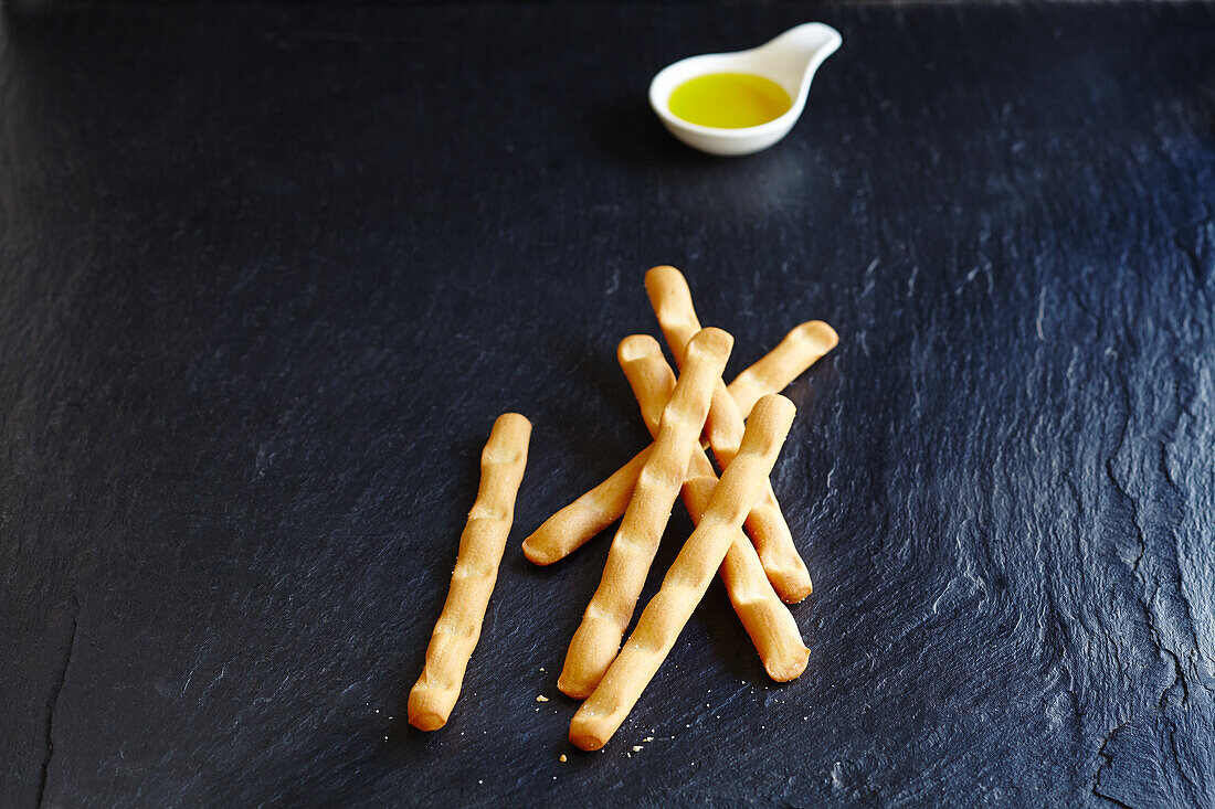 Breadsticks and a small bowl of olive oil on a slate surface