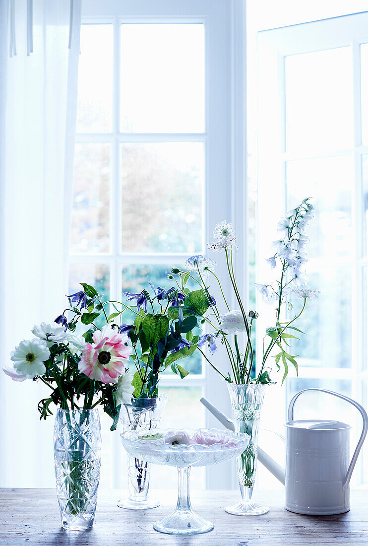 Spring flowers in glass vases, glass cake stand and watering can on table