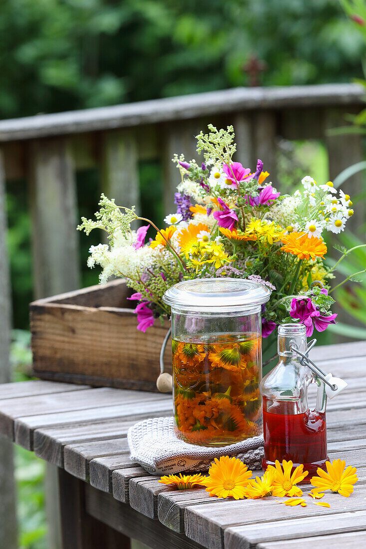 Kräuteröl, Sirup und Blütenstrauß auf Holztisch im Garten