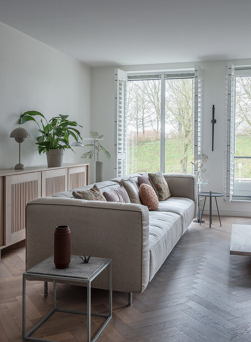 Modern living room with light grey sofa and cushions, wooden chest of drawers and large windows