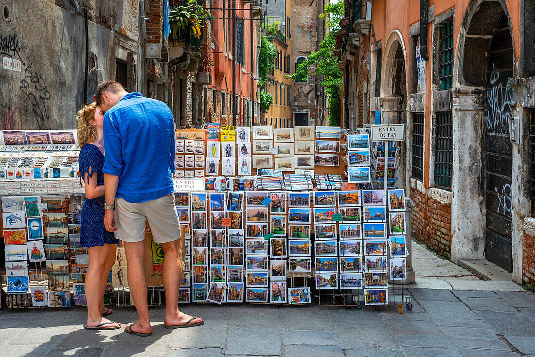 Romantisches Paar drückt seine Liebe aus, indem es sich im Stadtzentrum von Venedig neben einem Postkartengeschäft küsst. Venedig, Italien. Romantisches Paar küsst sich auf dem Gehweg des Kanals in der Nähe einer Brücke im historischen Teil der Stadt