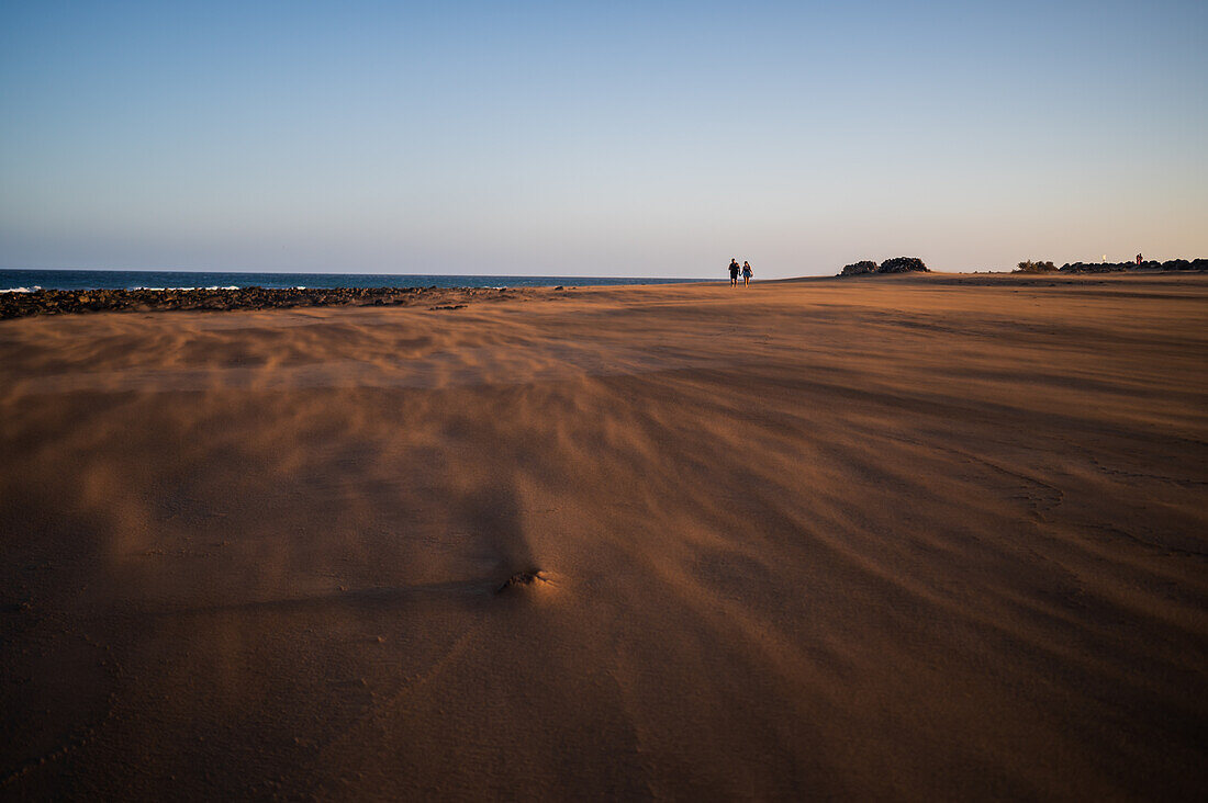 Ein Paar geht am Strand spazieren, während ein starker Wind den Sand auf Lanzarote, Kanarische Inseln, Spanien, weht